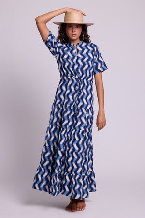A model wearing a KLEED short-sleeved, maxi dress with a blue and white wavy zigzag pattern, paired with a straw hat, against a solid light gray studio background.