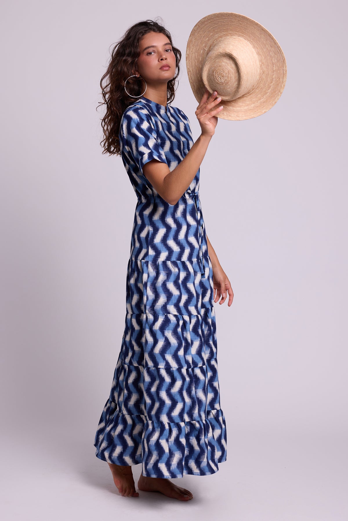 A model wearing a KLEED blue and white zigzag patterned maxi dress, holding a straw hat against a light gray studio background.