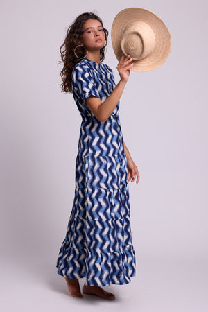 A model wearing a KLEED blue and white zigzag patterned maxi dress, holding a straw hat against a light gray studio background.