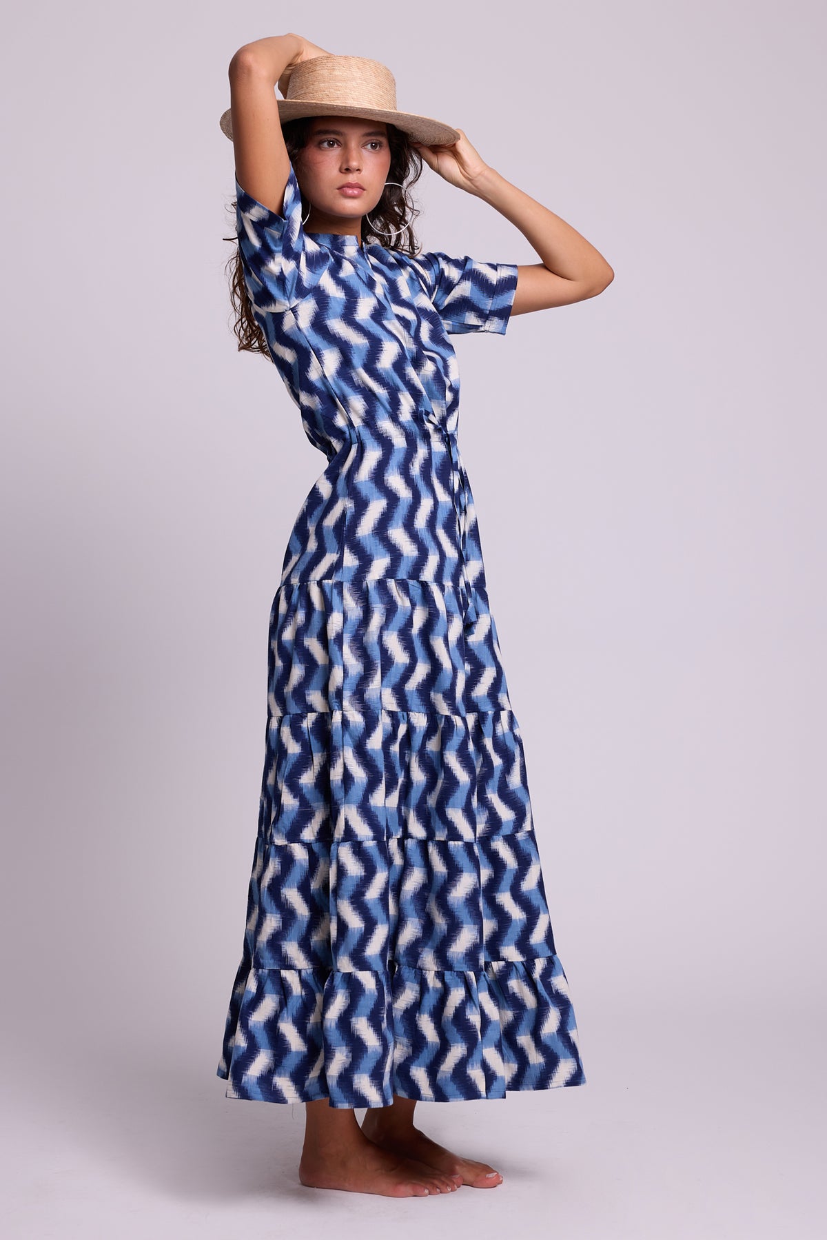 A woman modeling a KLEED short-sleeved, maxi dress with a blue and white wavy zigzag pattern, paired with a straw hat, against a solid light gray studio background.