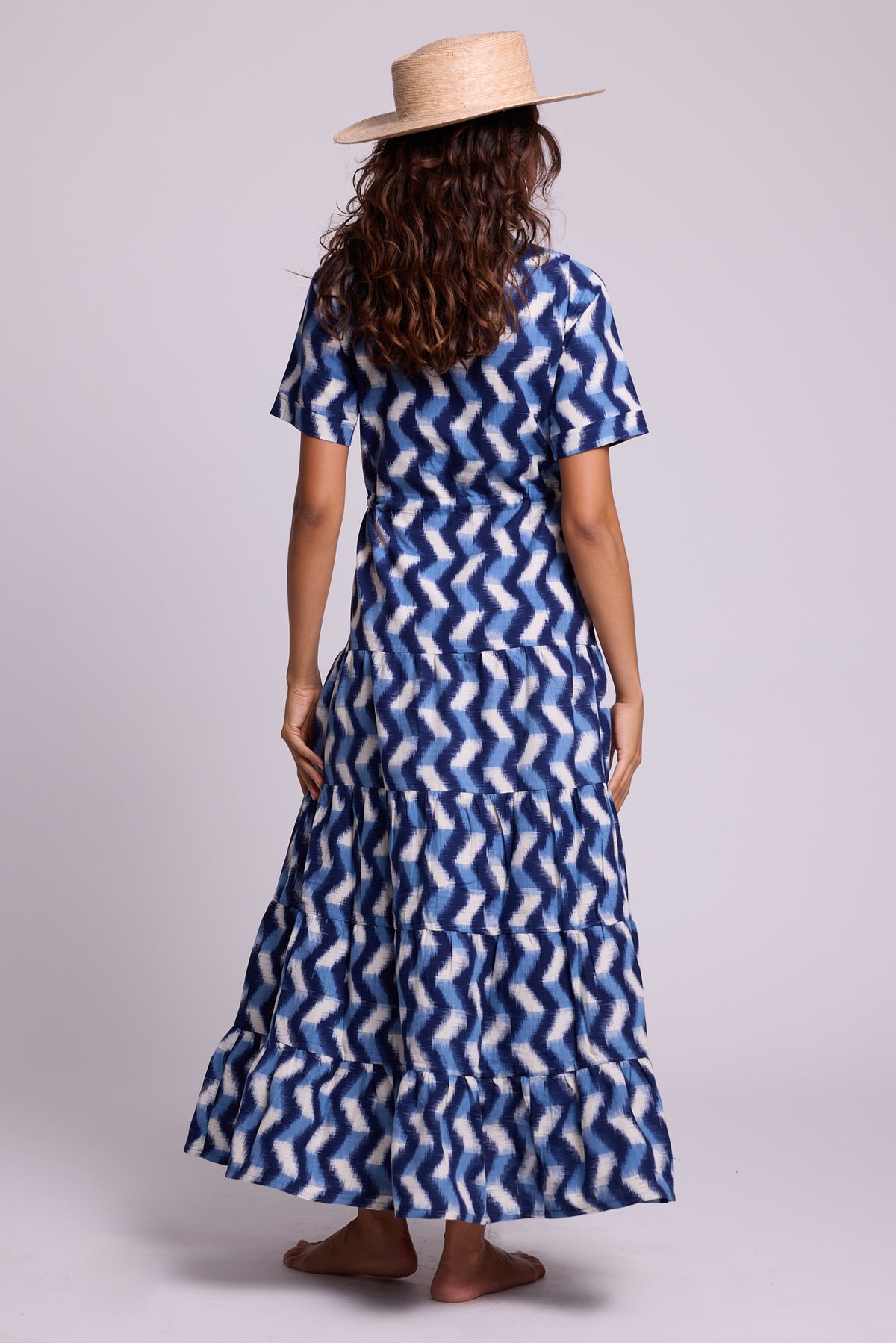 A model shown from the back wearing a KLEED short-sleeve, maxi dress with a blue and white pattern, paired with a straw hat, against a light gray studio background.
