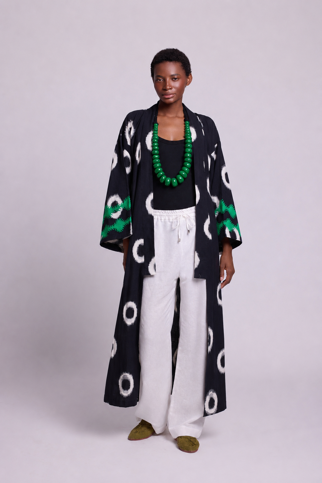 A model stands against a light gray studio background wearing a long black ikat kimono by kleed with white circular motifs and green patterned embroidereds  cuffs, over a black top and white wide-leg pants, accessorized with a chunky green beaded necklace and olive shoes.