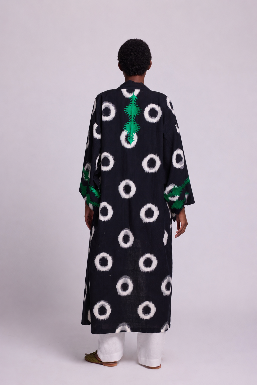 A model shown from the back wearing a cotton black ikat kimono from kleed with a white circular dot pattern and green embroidered details at the back and sleeves, paired with white pants, against a plain light gray studio background.