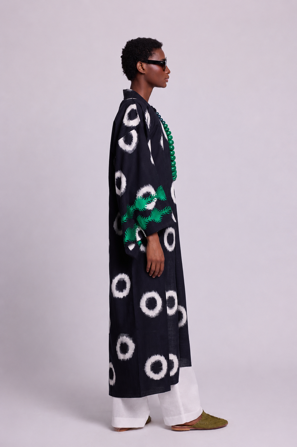 A model shown in profile wearing a long black ikat kleed kimono with circular patterns and an emerald green embroidery at the cuffs styled with white wide-leg pants and olive green shoes against a light gray studio background.