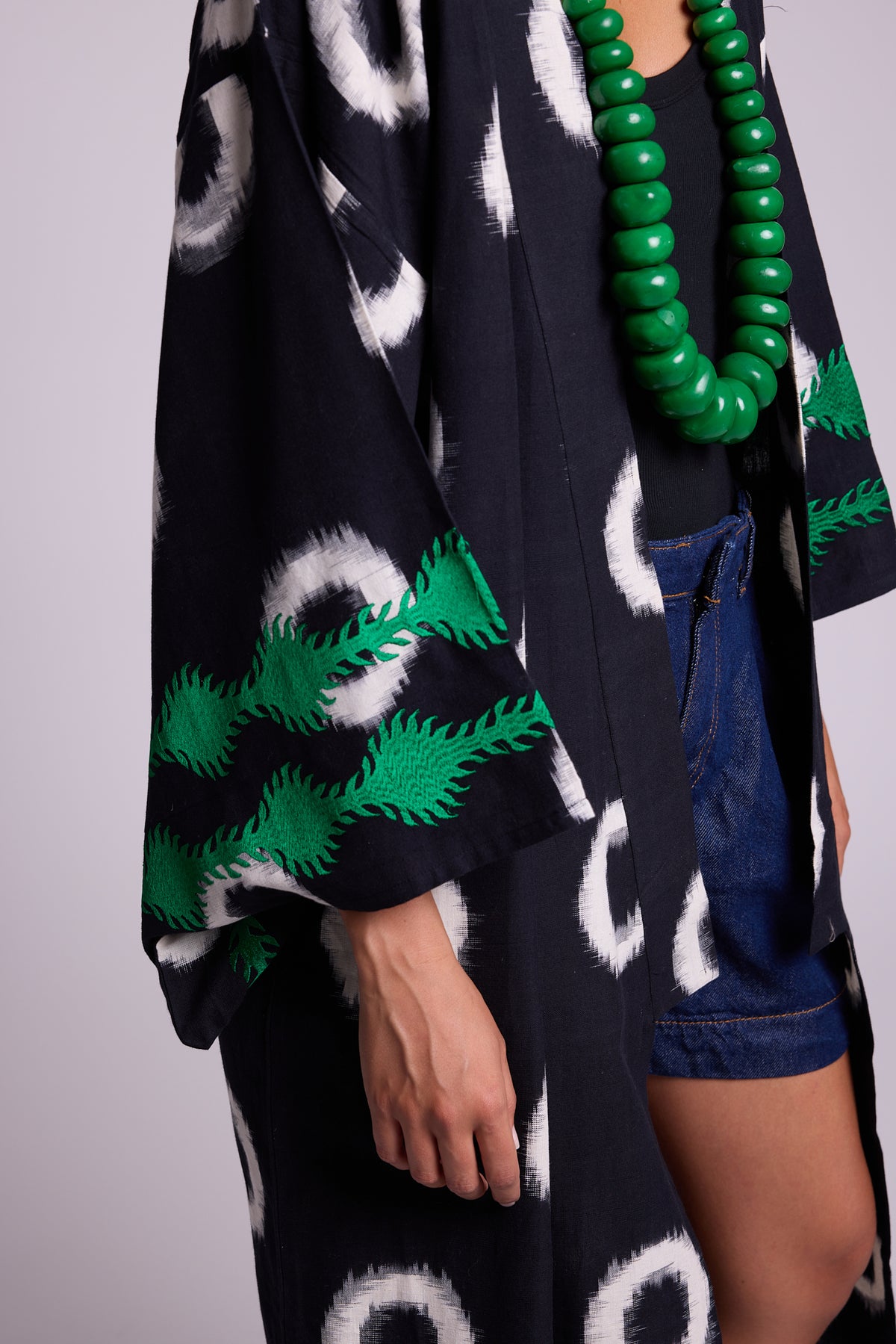 A close-up of a model wearing a black kimono- in cotton ikat by kleed, hand loomedm with white circular motifs and emerald green silk embroidery at the cuffs, styled with denim shorts, a chunky green beaded necklace, and a black top against a light gray studio background.