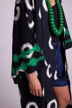 A close-up of a model wearing a black kimono- in cotton ikat by kleed, hand loomedm with white circular motifs and emerald green silk embroidery at the cuffs, styled with denim shorts, a chunky green beaded necklace, and a black top against a light gray studio background.