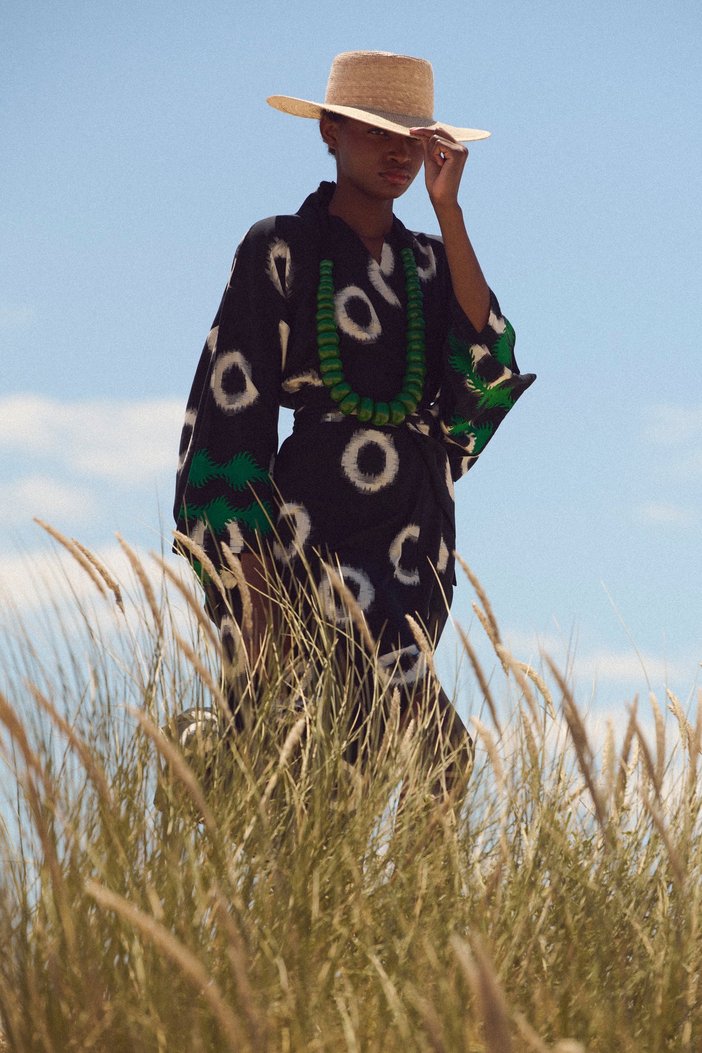 A model wearing a black ikart cotton kimono by kleed with white circular patterns, a matching straw hat, and a green beaded necklace, standing in tall grass under a blue sky.
