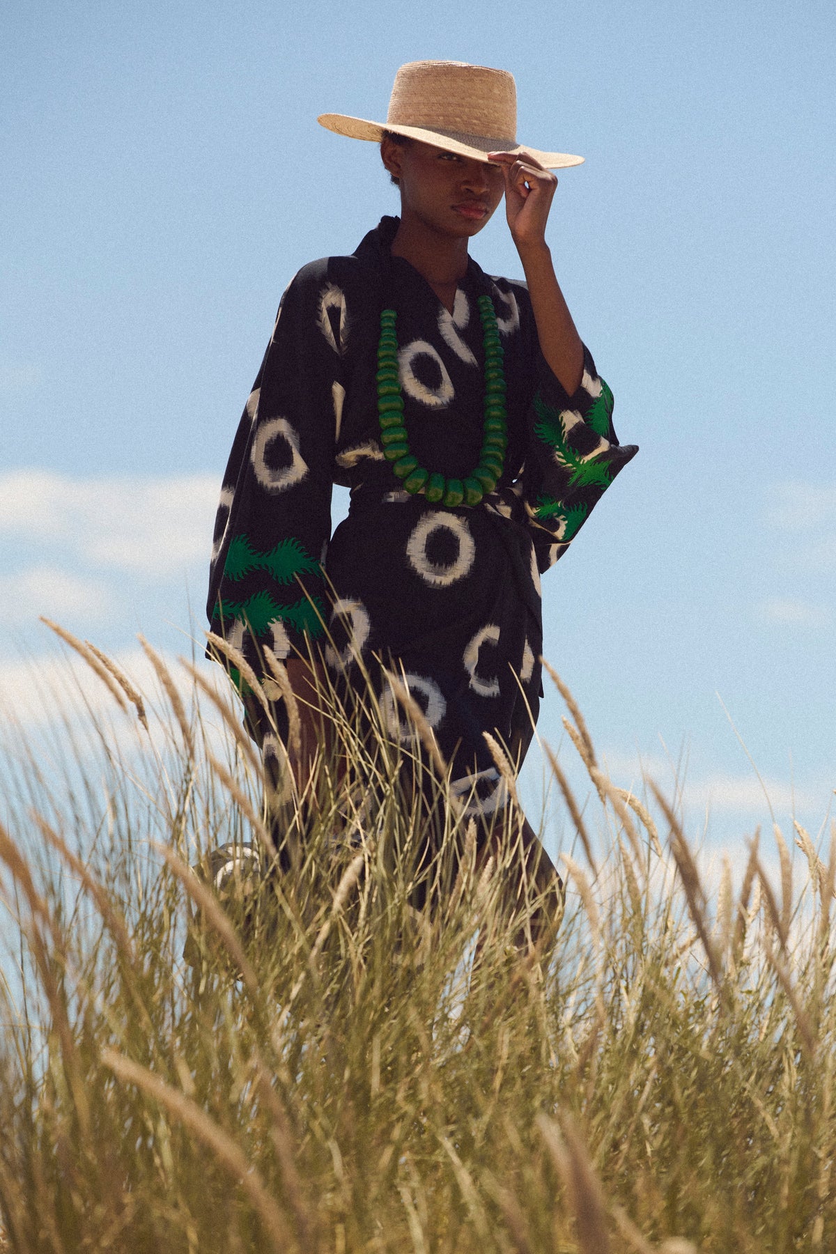 A model wearing a black ikart cotton kimono by kleed with white circular patterns, a matching straw hat, and a green beaded necklace, standing in tall grass under a blue sky.