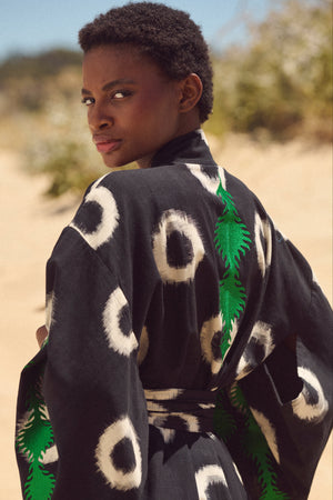 A model wearing a black kimono by kleed with white circular patterns and emerald green embroidery at the back, photographed outdoors in a sandy desert setting.