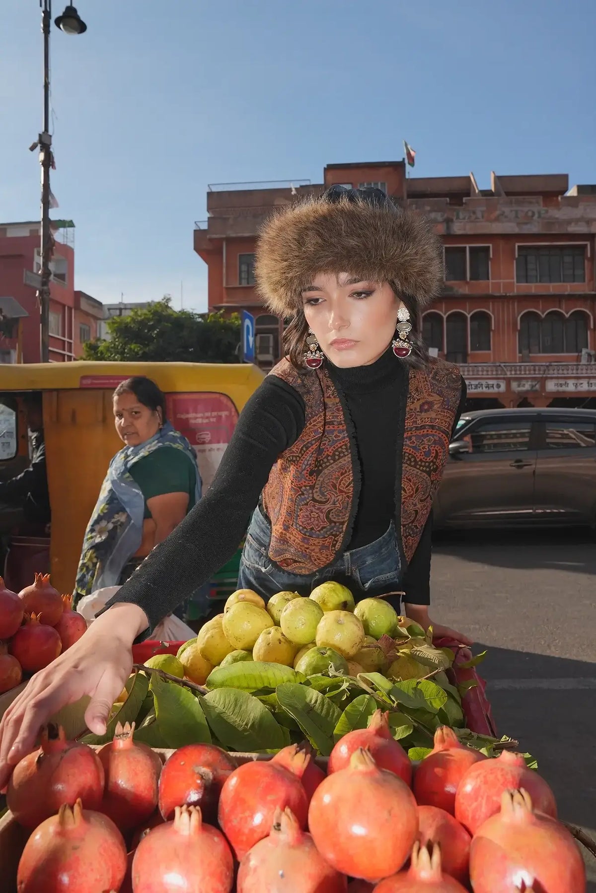 Model at a fruit market stall wearing Kleed Blue Oak Vest in wool with paisley print, surrounded by pomegranates and oranges.