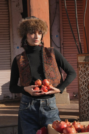 Model standing in a sunlit market wearing Kleed Blue Oak Vest in pure wool with paisley print, holding a tray of pomegranates.