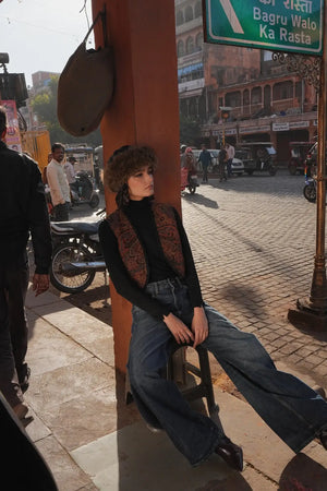 Model seated on a stool on a sunlit street wearing Kleed Blue Oak Vest in wool with paisley print, black turtleneck and wide-leg jeans.
