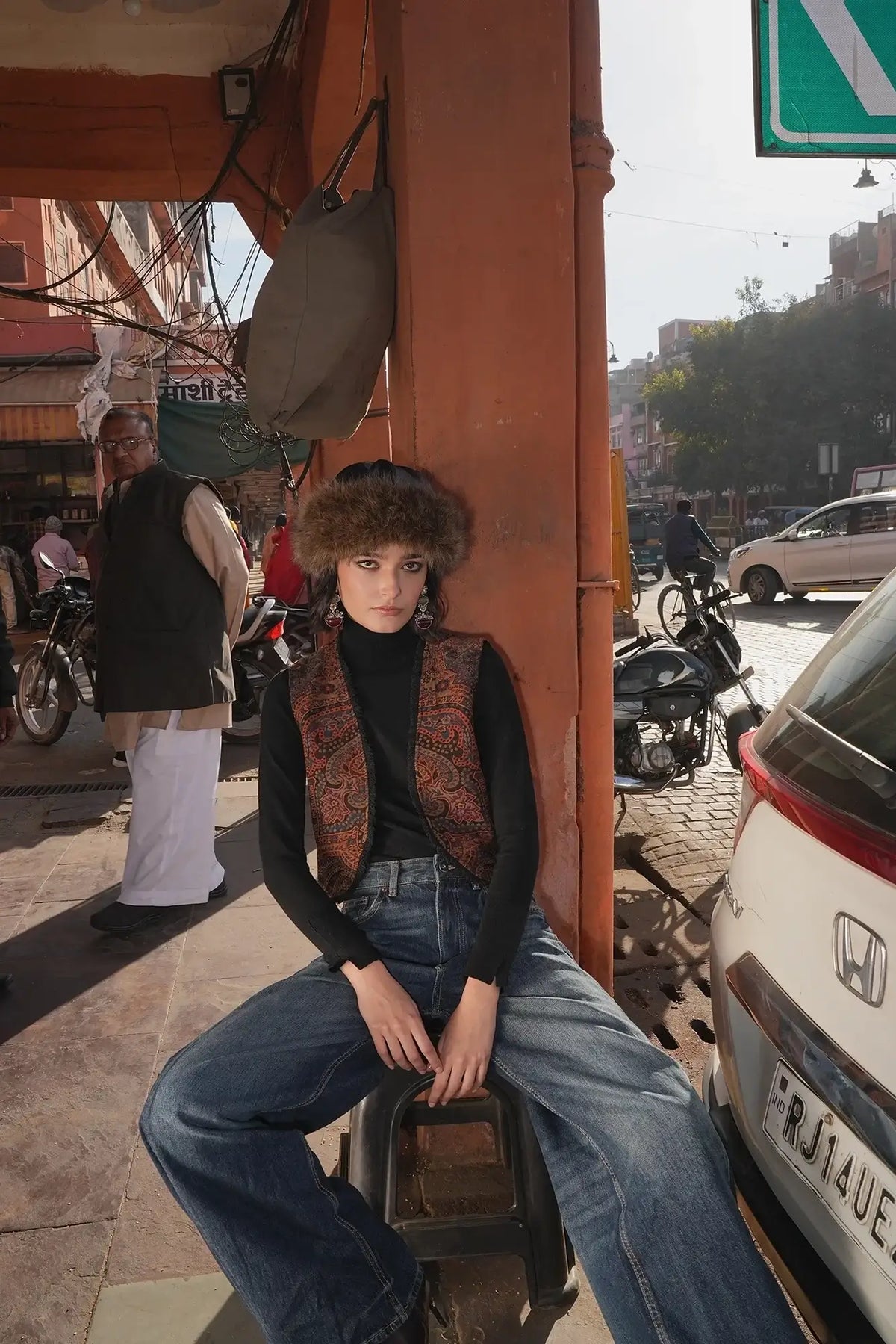Model seated on a low stool on a sunlit street wearing Kleed Blue Oak Vest in wool with paisley print, black turtleneck and wide-leg jeans.