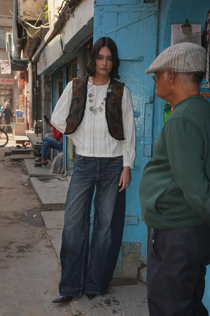 Model standing in front of a blue shopfront wearing Kleed Green Oak Vest in wool with paisley print, white blouse and jeans.