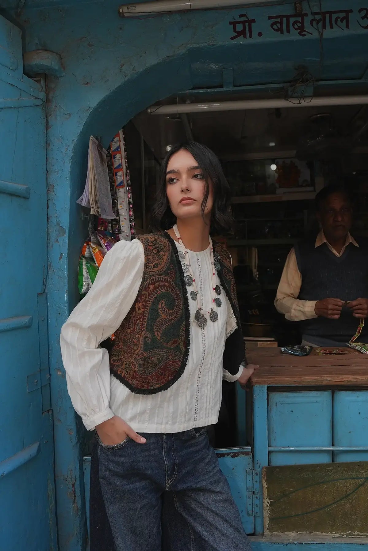 Model standing in front of a blue shopfront wearing Kleed Green Oak Vest in wool with paisley print, white blouse and jeans.