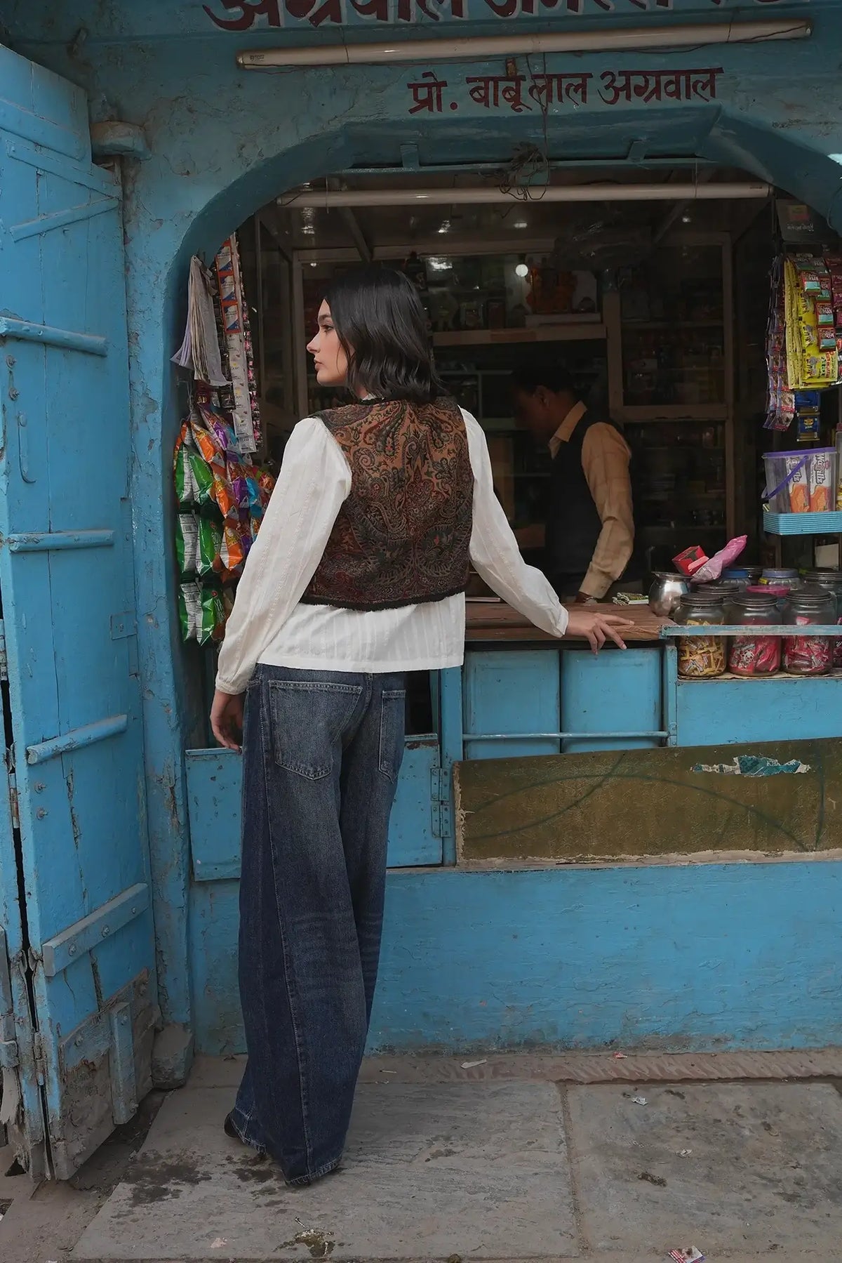 Model from behind on a blue painted street wearing Kleed Green Oak Vest in wool with paisley print and wide-leg jeans.