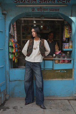 Model standing in front of a blue shopfront wearing Kleed Green Oak Vest in pure wool with paisley print, white top and wide-leg jeans.