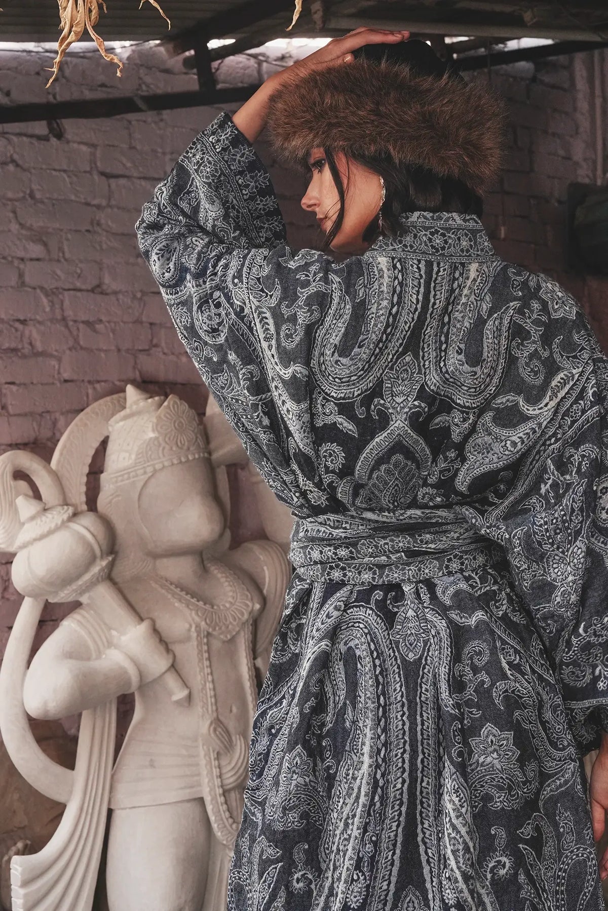 Back view of Kleed Blue Paisley Kimono Coat in pure wool with indigo paisley print, tied at the waist, editorial shot with a carved stone sculpture.