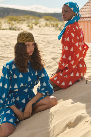 Two models seated on a sandy beach wearing Kleed Turquoise Savannah Short Kimono in handwoven Ikat cotton with white triangle print and orange savannha short kimono.
