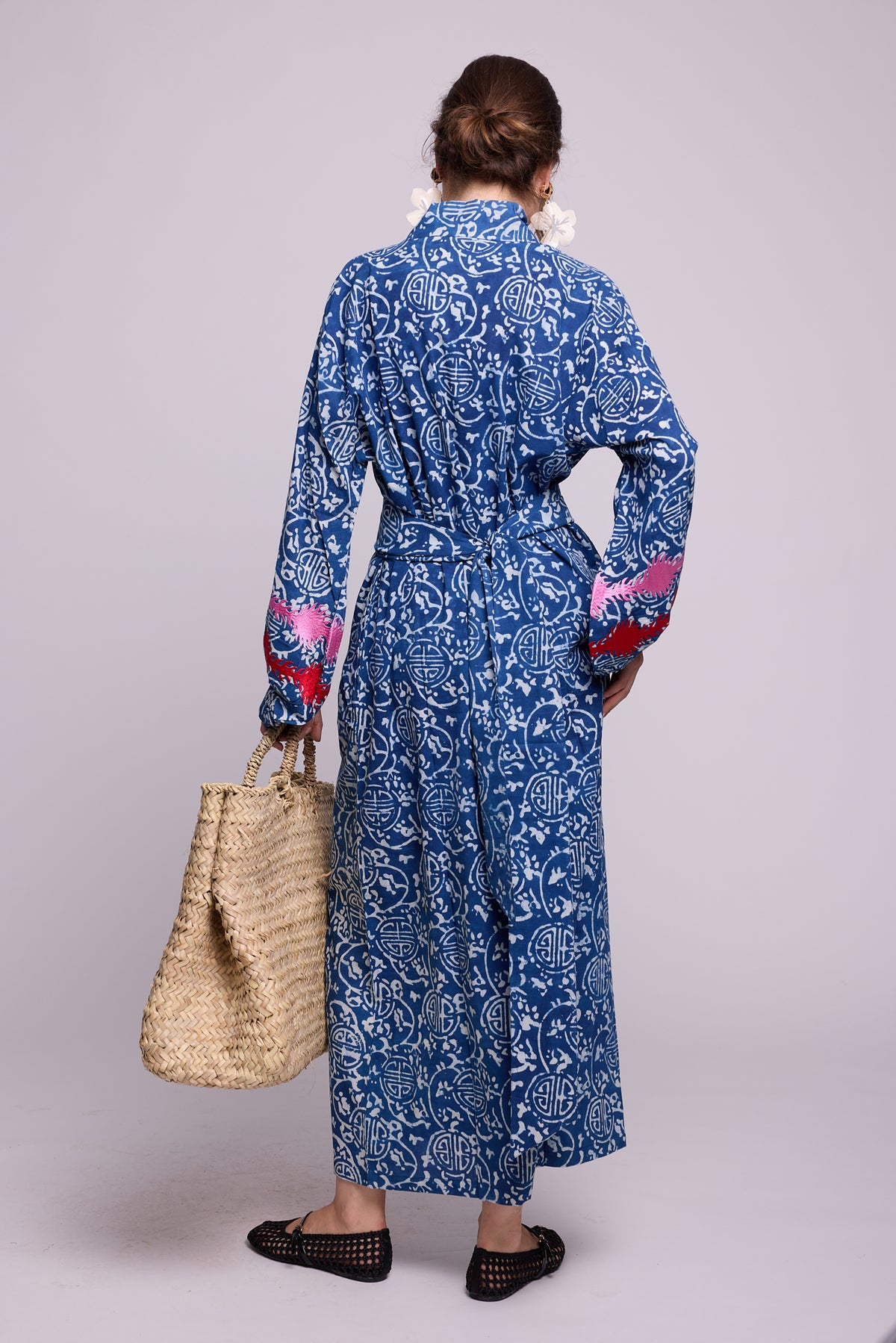 Back view of a woman wearing KLEED’s 100% chemical-free, hand block-printed Indira Bloom Kimono in indigo blue and white, featuring pink and red embroidered balloon sleeves, a matching belt and a straw tote bag.
