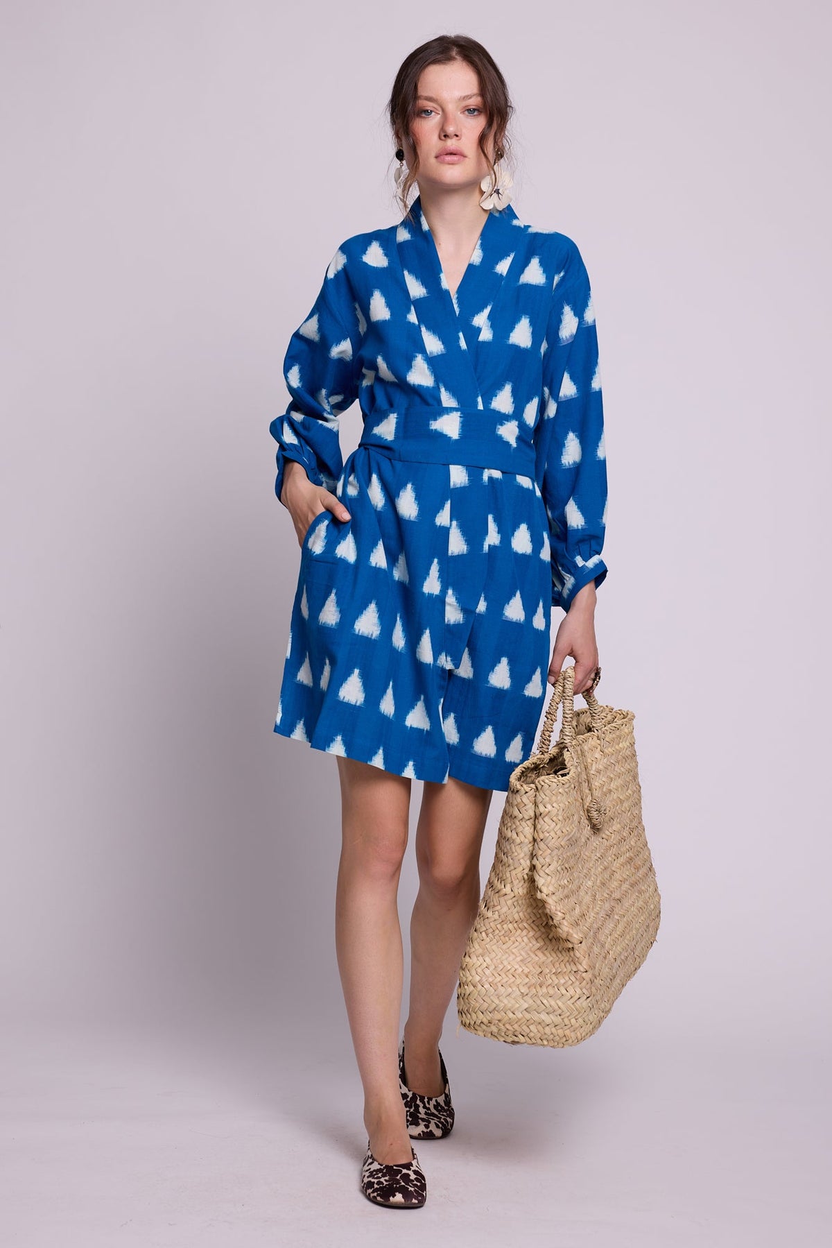 Model standing wearing Kleed Turquoise Savannah Short Kimono in Ikat cotton with white triangle print, holding a straw tote.