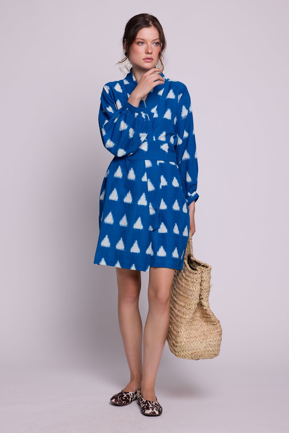 Model standing wearing Kleed Turquoise Savannah Short Kimono in handwoven Ikat cotton with white triangle print, styled with a straw tote and animal print mules.