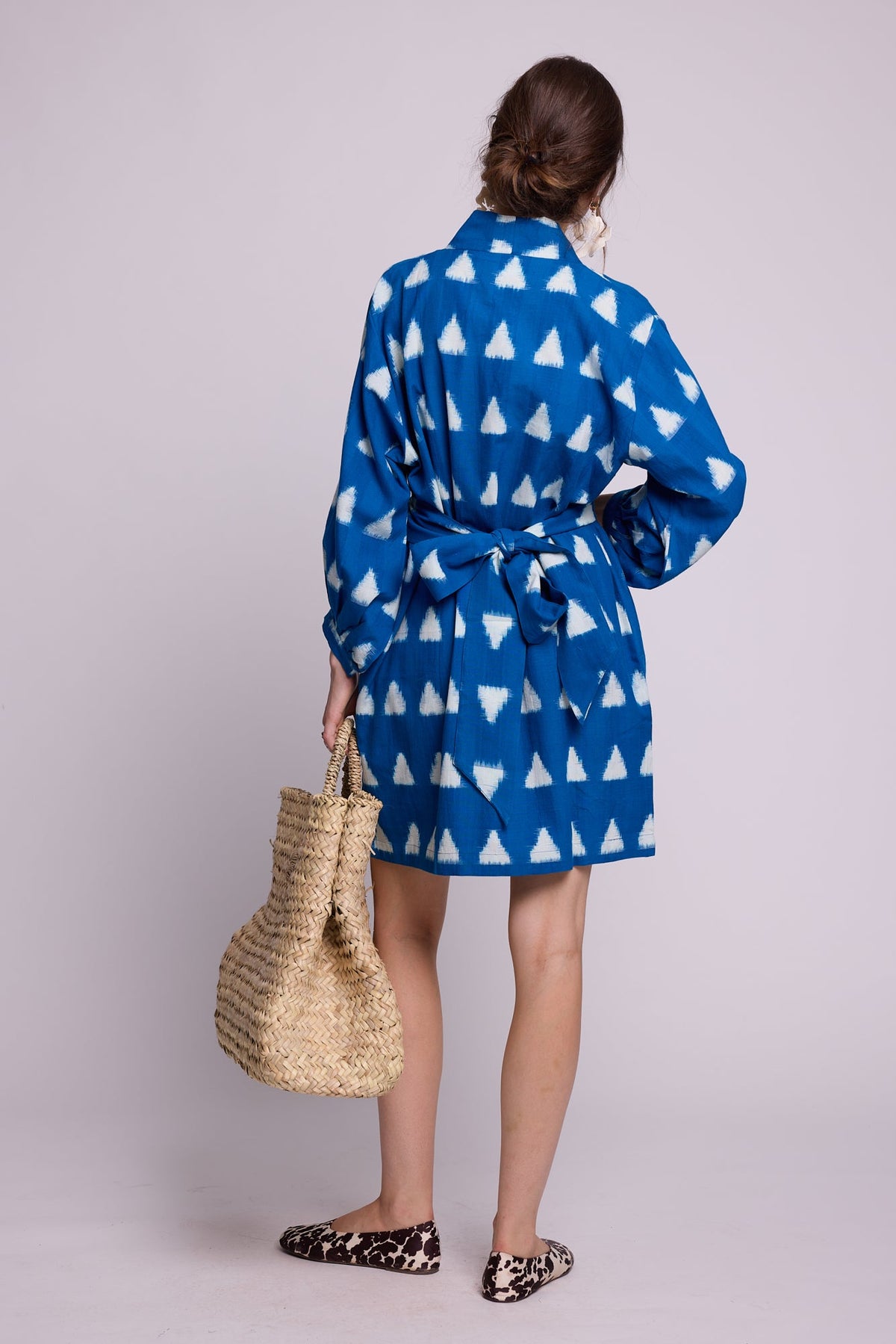 Model from behind wearing Kleed Turquoise Savannah Short Kimono in Ikat cotton with white triangle print, holding a straw tote.