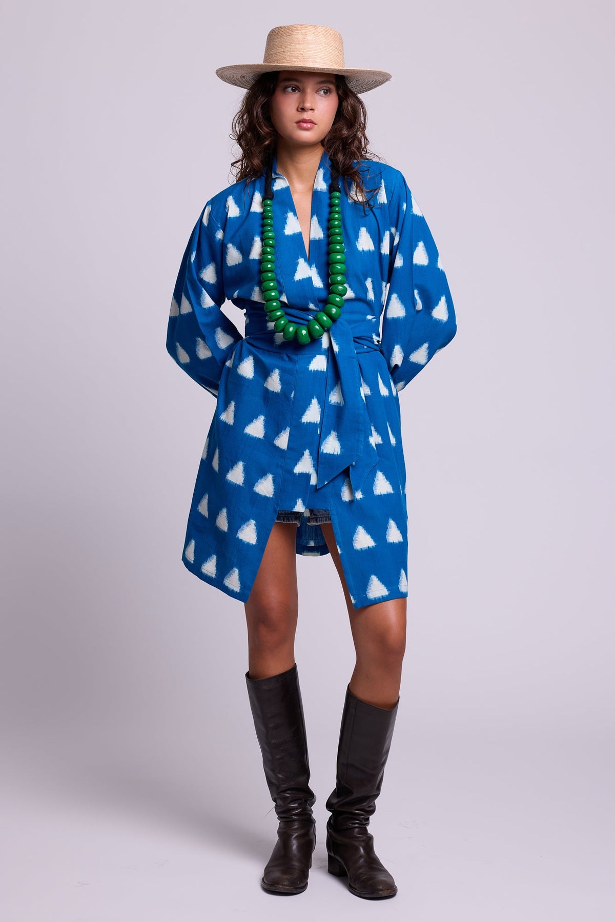 Model seated on a sandy beach wearing Kleed Turquoise Savannah Short Kimono in handwoven Ikat cotton with a straw hat.

