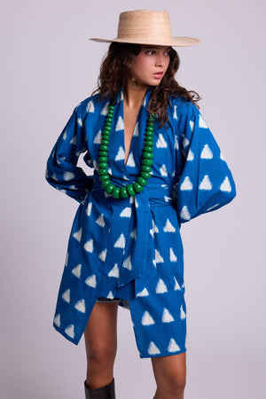 Model standing wearing Kleed Turquoise Savannah Short Kimono in handwoven Ikat cotton with white triangle print, styled with a straw hat and green beaded necklace.