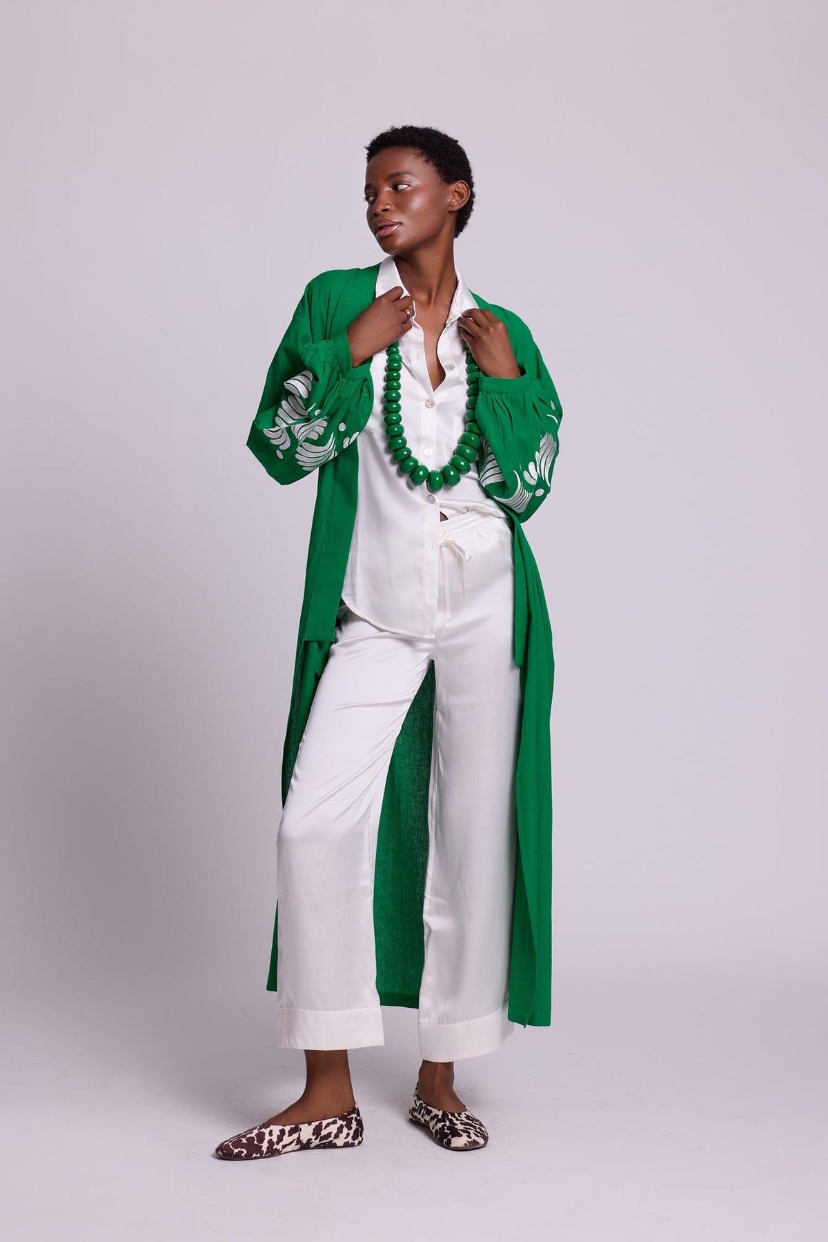 Full length view of a woman wearing the Kleed Green Kandy Kimono in 100% cotton with white floral embroidered sleeves, styled with white pyjama, green beaded necklace and leopard print flat mules