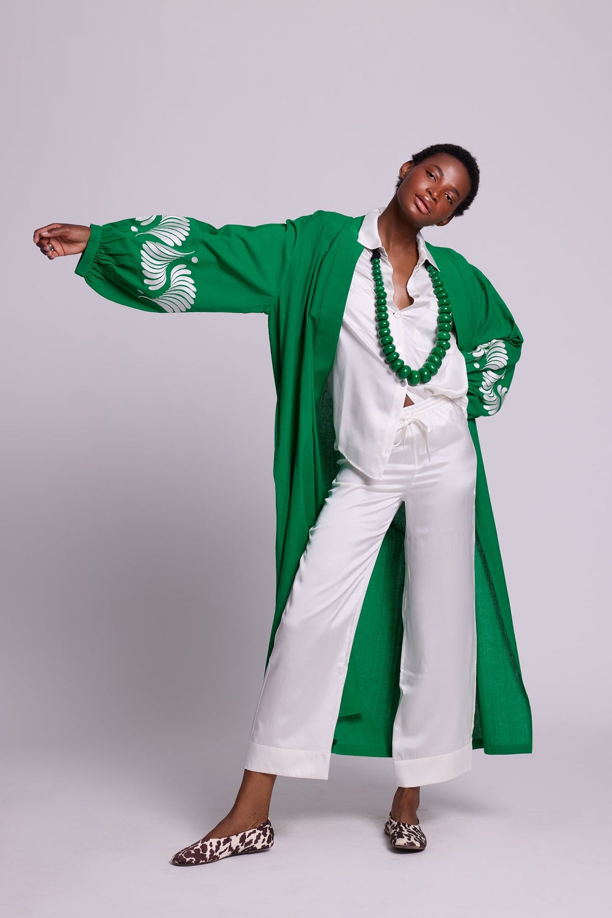 Woman wearing Kleed´s Green Kandy Kimono in 100% cotton with white floral embroidered sleeves, arms outstretched, styled with white silk pyjama, green beaded necklace and leopard print flat mules