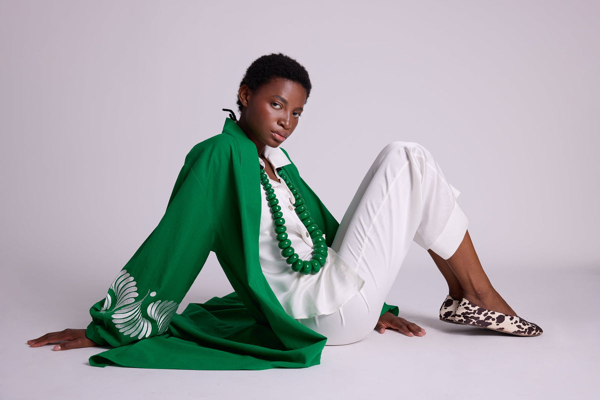 Woman seated on the floor wearing the Kleed Green Kandy Kimono in 100% cotton with white floral embroidered sleeves, styled with white silk pyjamas and green beaded necklace