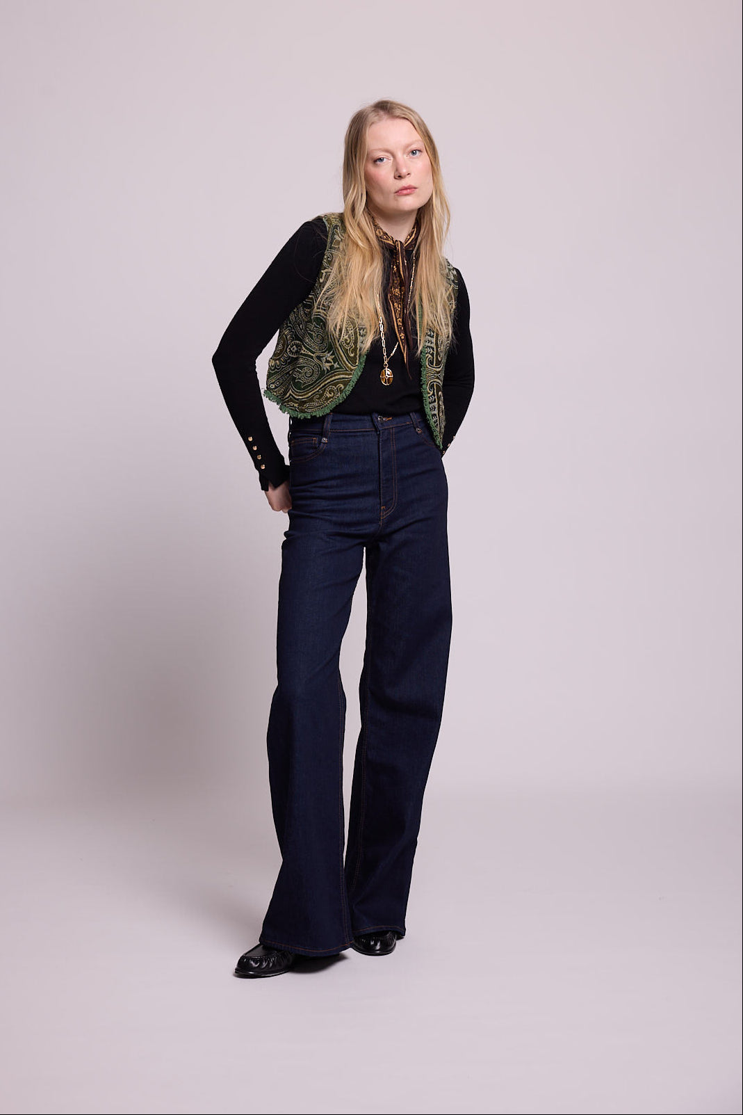 Full length view of a woman wearing the Kleed Green Paisley Wool Vest over a black top and dark wide leg jeans, alternative pose