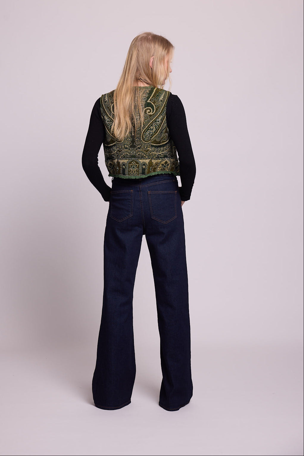 Full length bacj view of a woman wearing the Kleed Green Paisley Wool Vest over a black long sleeve top and dark wide leg jeans
