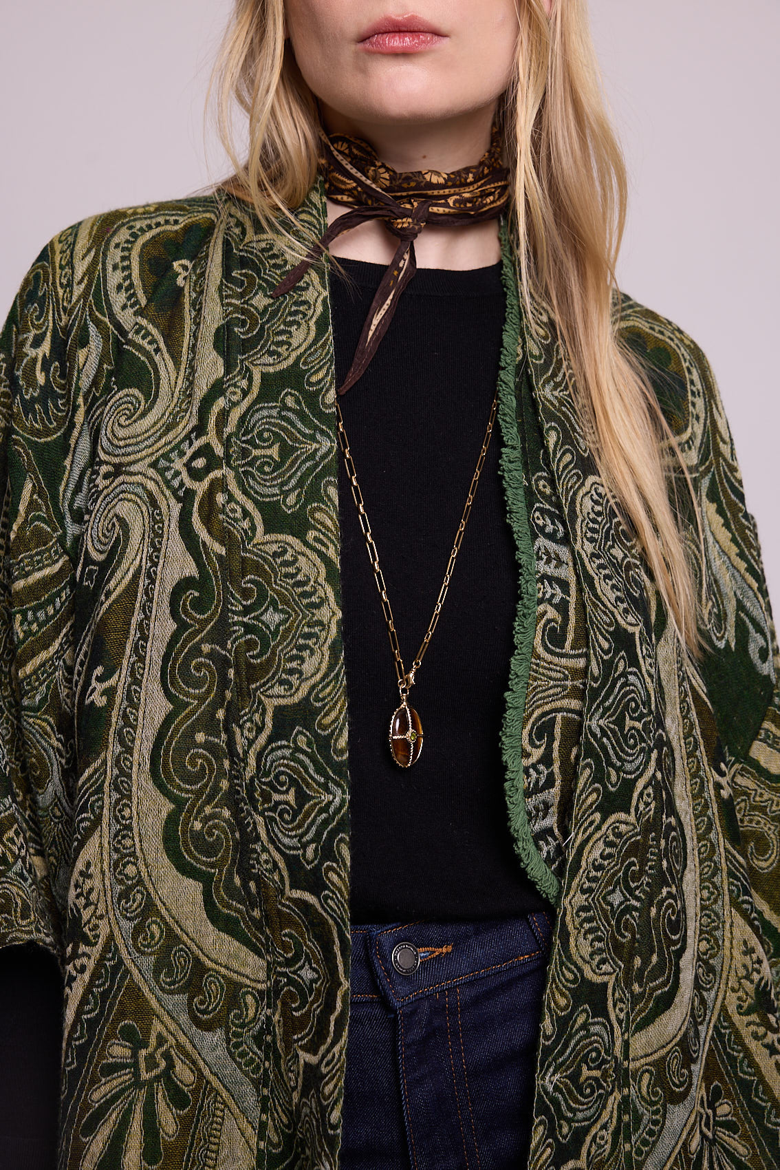 Close up detail of a woman wearing the Kleed Green Paisley Wool Vest layered under a green paisley kimono coat, styled with a black ribbed top, dark jeans, a printed neck scarf and a gold oval pendant necklace