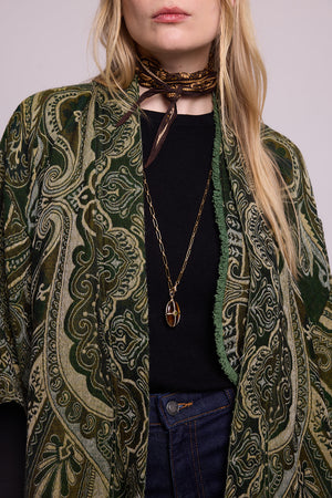 Close up detail of a woman wearing the Kleed Green Paisley Wool Vest layered under a green paisley kimono coat, styled with a black ribbed top, dark jeans, a printed neck scarf and a gold oval pendant necklace