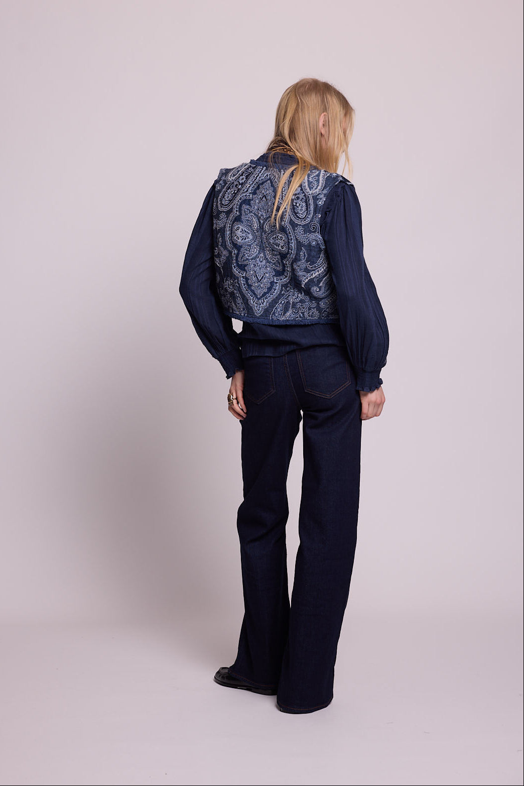 Back view of a woman wearing the Kleed Blue Paisley Vest showing the intricate handcrafted paisley pattern, styled with a navy blouse and dark wide leg jeans