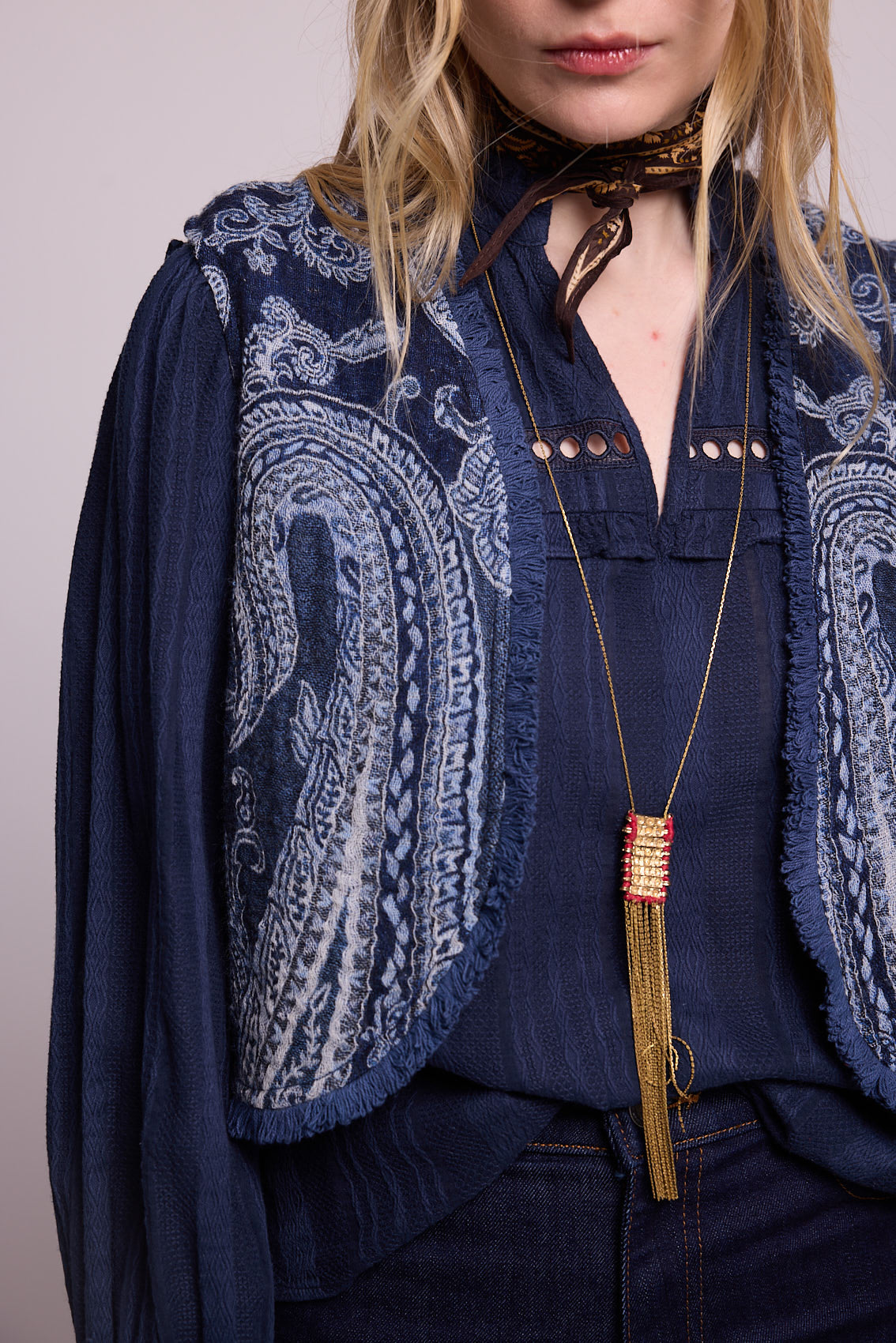 Close up detail of the Kleed Blue Paisley Vest showing the handcrafted paisley pattern, fringe trim edges and fabric texture, styled with a gold tassel pendant necklace and dark neck scarf