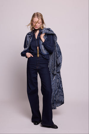 Full length view of a woman wearing the Kleed Blue Paisley Vest over a navy blouse and dark wide leg jeans, holding a blue paisley kimono coat draped over her shoulder, styled with a dark neck scarf and gold necklace