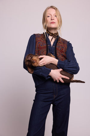 Model standing wearing Kleed Blue Oak Vest in pure wool with paisley print, navy blouse and dark jeans, holding a dachshund.