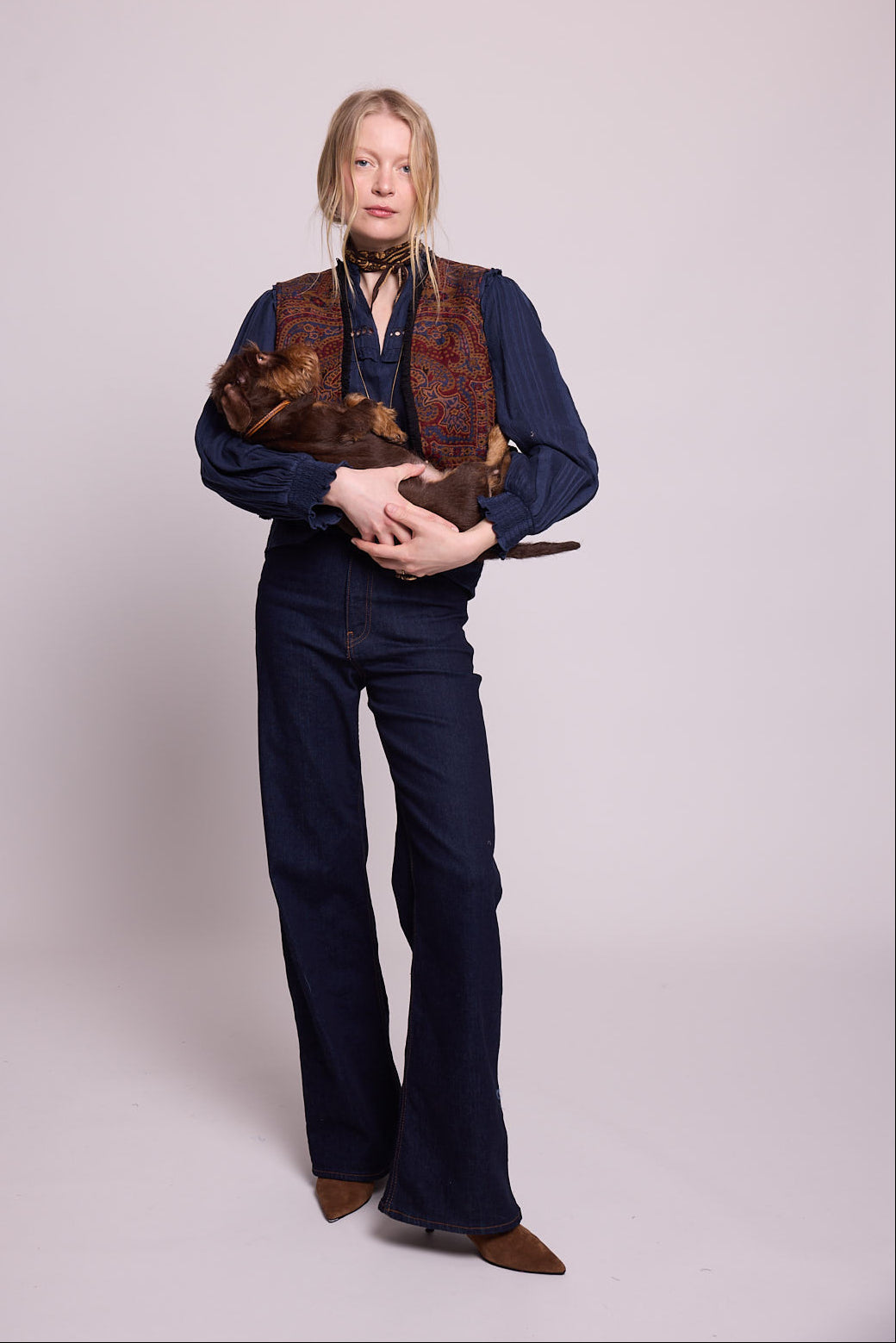 Model standing wearing Kleed Blue Oak Vest in wool with paisley print, navy balloon-sleeve blouse and dark wide-leg jeans, holding a dachshund.