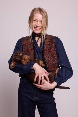 Model smiling in studio wearing Kleed Blue Oak Vest in pure wool with paisley print, navy balloon-sleeve blouse and silk scarf, holding a wirehaired dachshund.