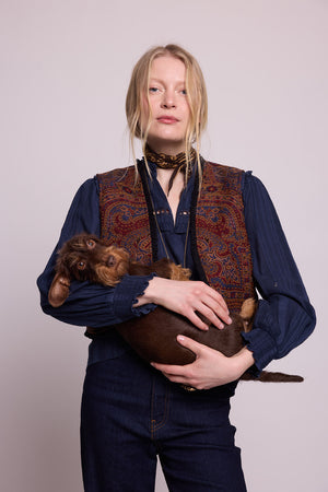Model standing in studio wearing Kleed Blue Oak Vest in pure wool with paisley print, navy blouse and silk scarf, holding a wirehaired dachshund.
