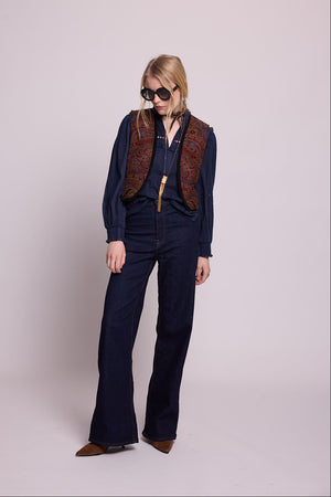 Model standing wearing Kleed Blue Oak Vest in wool with paisley print, navy balloon-sleeve blouse and dark wide-leg jeans, holding a dachshund.