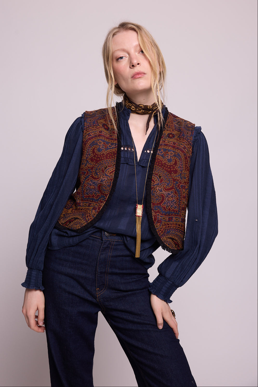 Model standing wearing Kleed Blue Oak Vest in pure wool with paisley print and fringe trim, navy blouse, silk scarf and gold pendant necklace.