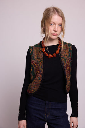Model standing wearing Kleed Green Oak Vest in pure wool with forest green and burgundy paisley print, over a black top with coral necklace.
