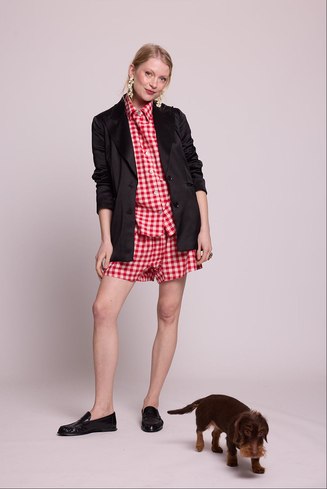 Full length view of a woman wearing the Kleed Bhati Red Pyjama Set in red vichy cotton styled with a black blazer and black loafers, with a small dachshund dog at her feet