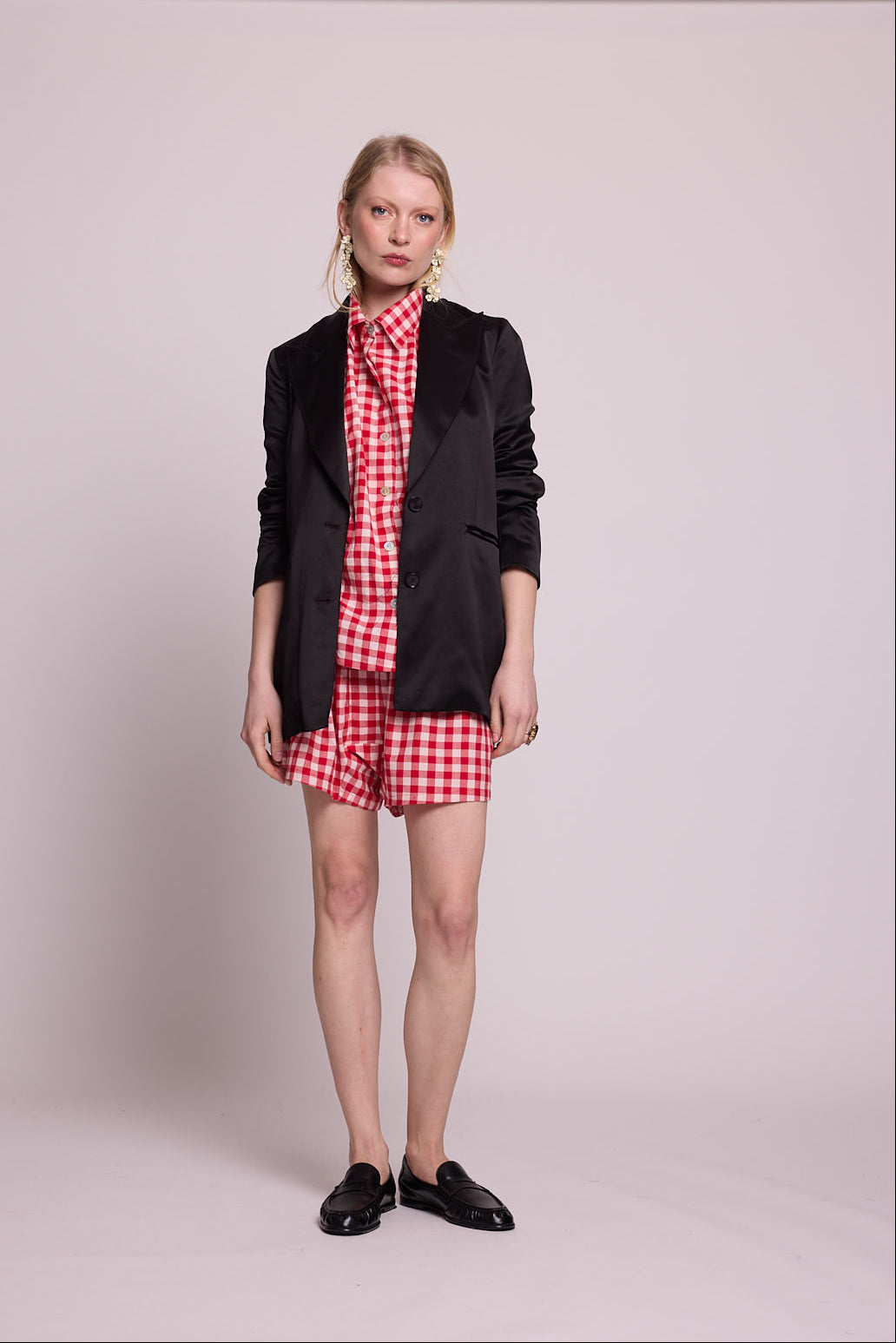 Full length front view of a woman wearing the Kleed Bhati Red Pyjama Set in red vichy cotton styled with a black blazer, black loafers and gold statement earrings, neutral expression