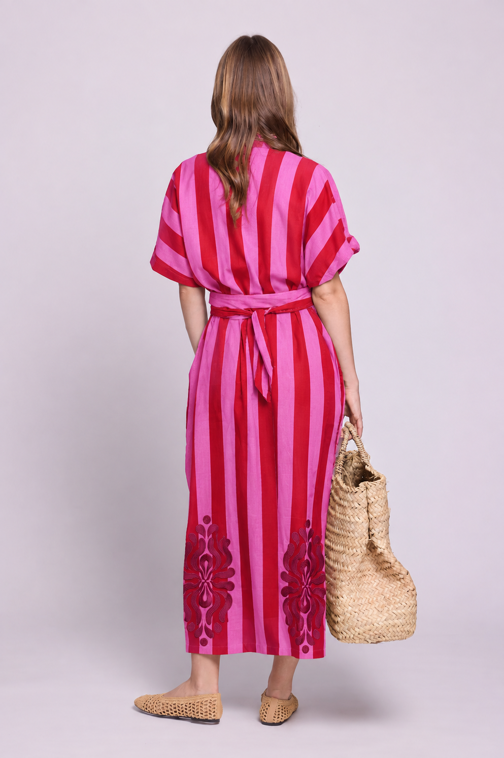 Rear view of a model wearing the Kleed Strawberries Kaftan, crafted in 100% cotton with pink and red hand block printed stripes, silk embroidery along the hem, side pockets and a matching waist belt, styled with an oversized woven straw bag and flat woven shoes.