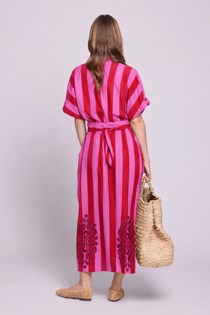 Rear view of a model wearing the Kleed Strawberries Kaftan, crafted in 100% cotton with pink and red hand block printed stripes, silk embroidery along the hem, side pockets and a matching waist belt, styled with an oversized woven straw bag and flat woven shoes.